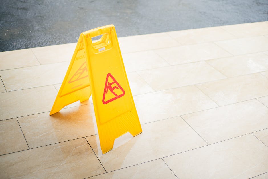 A yellow caution wet floor sign with red hazard symbols is positioned on a beige ceramic tiled floor in an indoor setting, indicating recent cleaning or maintenance work. The tiles are clean, shiny, and appear to be well-maintained, reflecting the ambient light. The sign is placed near the edge of the tiled area, suggesting a need for caution to prevent slipping. The surrounding area is smooth, with visible grout lines between the tiles, and the lighting highlights the cleanliness and orderliness of the space, consistent with professional surface cleaning and sanitisation services provided by Cleaners W6, aligned with the aim of avoiding hidden cleaning charges in Hammersmith bookings, W6.