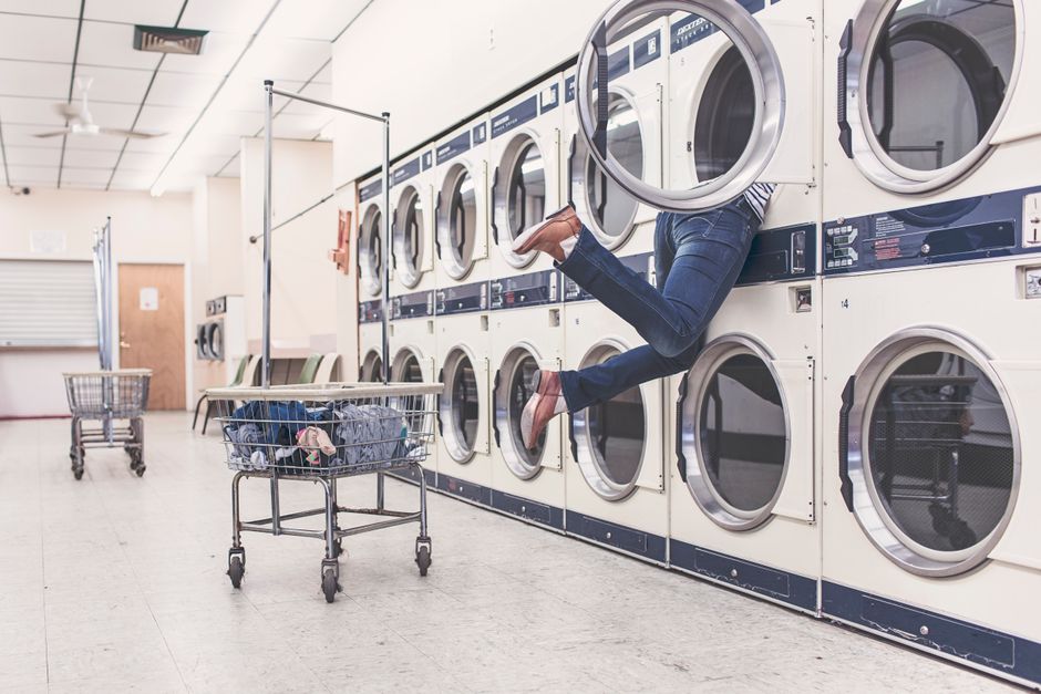 An individual with legs wearing blue jeans and brown shoes is leaning out of a washing machine door in a laundromat, with their upper body obscured inside the machine. The setting features a row of front-loading washing machines with silver doors and control panels, set against a white tiled wall. A shopping cart filled with clothes and laundry supplies is positioned in front of the machines. The laundromat has overhead lighting, a light-colored tiled floor, and a wooden door visible in the background. Cleaners W6 provides comprehensive domestic cleaning, including surface cleaning and sanitisation, to address common laundry and hygiene issues in residential spaces near Hammersmith, W6.