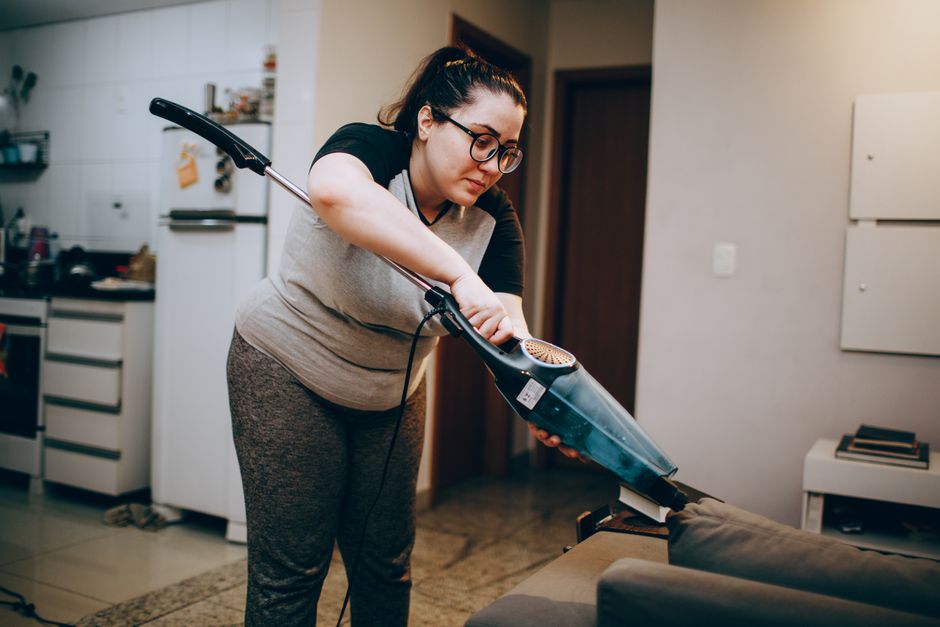 A woman wearing glasses and casual clothing is engaged in domestic surface cleaning, using a handheld vacuum cleaner to meticulously vacuum a sofa in a living room. The room features a beige carpet, a dark-colored sofa with cushions, and light-colored walls with minimal decorations. Visible in the background are a kitchen area with white cabinets, a refrigerator, and various household items. Sunlight or indoor lighting illuminates the space, highlighting the clean and well-maintained surfaces. This scene exemplifies detailed deep cleaning and sanitisation practices, as part of professional household cleaning services provided by Cleaners W6, aimed at maintaining hygiene and addressing common apartment cleaning challenges in Hammersmith, W6.