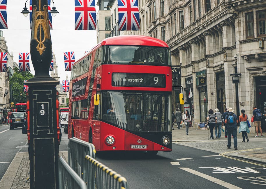 A red double-decker bus with the route number 9 and destination Hammersmith traveling along a busy city street lined with historic buildings. British Union Jack flags are hanging from street lamps, and pedestrians are walking along the sidewalk. The scene is outdoors during daytime with overcast weather, capturing the urban atmosphere typical of central London. Cleaners W6 offers professional cleaning services for residential and commercial properties, including surface cleaning, deep cleaning, and sanitisation, ensuring spaces are maintained to the highest hygiene standards.