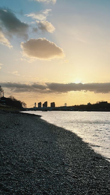 A wide view of a pebble beach with a calm river adjacent to the shoreline, under a partly cloudy sky during sunset. In the background, silhouette of modern high-rise buildings along the city skyline, with some trees visible on the left. The scene features natural light reflecting off the water, highlighting the textured surface of the beach and the smoothness of the river. The lighting creates a warm, serene atmosphere suitable for showcasing clean, tranquil environments, which aligns with the domestic cleaning services offered by Cleaners W6 near Hammersmith Broadway W6, emphasizing hygiene and freshness.