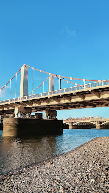 The image features a suspension bridge spanning a river, with large concrete supports and metal cables holding the structure. The bridge has a white and red painted railing and a walkway beneath the main deck. The river below is calm, with a pebble-covered shoreline in the foreground. Bright, clear blue sky with minimal clouds provides natural lighting, highlighting the clean surfaces of the bridge and surroundings. This scene reflects the importance of maintaining structural cleanliness and surface safety for bridges in riverside flats in Hammersmith. For comprehensive surface cleaning and deep sanitisation of similar outdoor infrastructure, [COMPANY_NAME], as featured on the Hammersmith Bridge cleaning tips page for riverside flats, offers expert services tailored to preserving the integrity and appearance of such structures within the W6 area.
