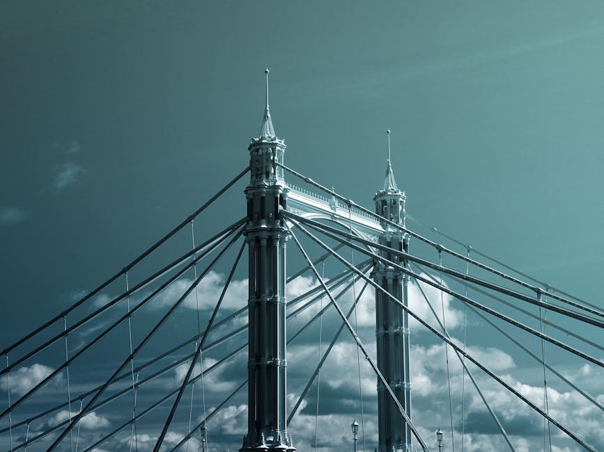 A detailed view of the suspension bridges of Hammersmith Bridge, featuring two tall, ornate towers with decorative spires, connected by numerous steel cables that fan out from the top of each tower. The bridge's dark metal structure contrasts with the partly cloudy sky in the background, highlighting its architectural features and the intricate cable arrangement. The image captures the bridge's historical design and grandeur against a natural sky scene, emphasizing the importance of surface and structural maintenance for heritage-listed structures, as discussed in cleaning and preservation tips for riverside flats on Cleaners W6's website.