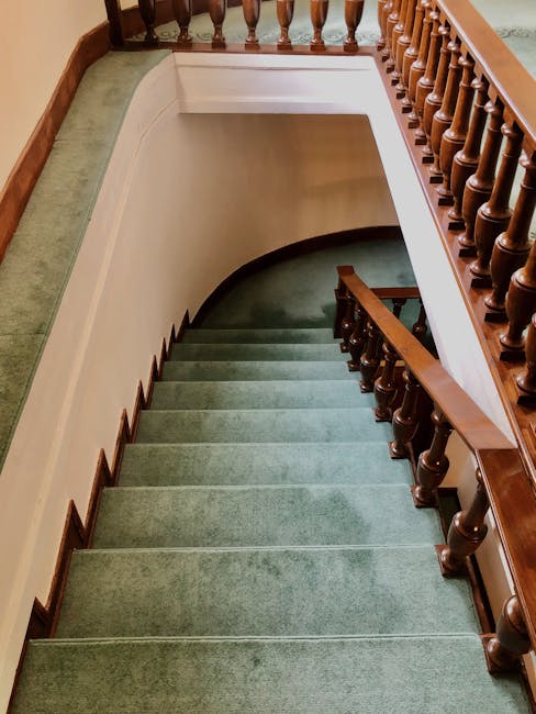 Interior view of a staircase with green carpeted steps, wooden handrail, and spindles, leading down to a lower floor, with clean and well-maintained surfaces. The staircase is illuminated by natural light, highlighting the tidy and polished appearance of the carpet and woodwork. The surrounding walls are painted in a light, neutral color, and a green carpet runner runs along the top landing area. The scene reflects professional surface cleaning and maintenance, as carried out by Cleaners W6, specialists in domestic cleaning for the Hammersmith, W6 area.
