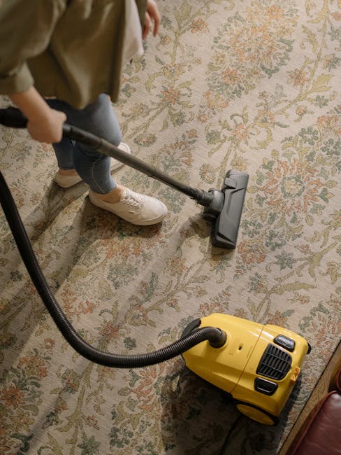 A person vacuuming a large, patterned carpet in a residential room with a yellow vacuum cleaner. The individual is wearing casual clothing, including beige shoes and jeans, and is holding a vacuum hose attachment directing it across the carpet's surface. The carpet features a floral design with muted colors, and the room appears tidy and well-maintained. The scene highlights surface cleaning activity typical of domestic cleaning services, with focus on deep cleaning and sanitisation facilitated by professional equipment, as promoted by Cleaners W6 on their King Street carpet cleaning services page in Hammersmith, W6.