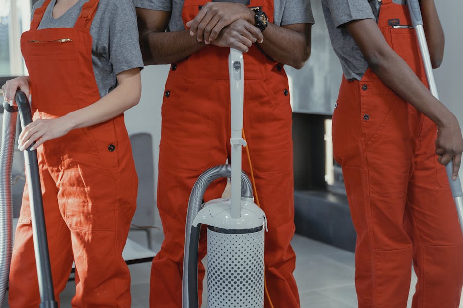 Close-up view of three commercial cleaners dressed in grey shirts and bright orange overalls, holding vacuum cleaners and cleaning tools, inside a well-lit, modern office space with a tiled floor and