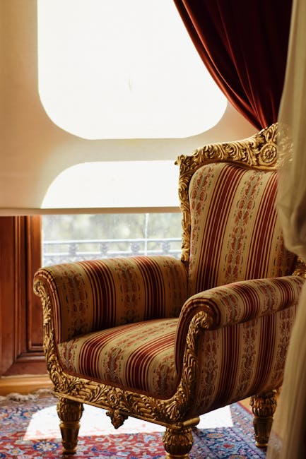 An ornate, upholstered armchair with carved wooden legs and decorative trim sits in front of a large, diffused window with a white semi-circular top, allowing natural light to fill the room. The fabric features a traditional pattern with red, gold, and beige tones. To the right, a deep red velvet curtain partially covers the window, adding a rich texture to the setting. The floor beneath the chair is covered with a patterned rug, and part of a wooden furniture piece is visible on the left side. The overall scene highlights a clean, well-maintained interior in a residential space, fitting for deep cleaning or surface care by Cleaners W6, who specialise in upholstery cleaning as part of their domestic cleaning services.
