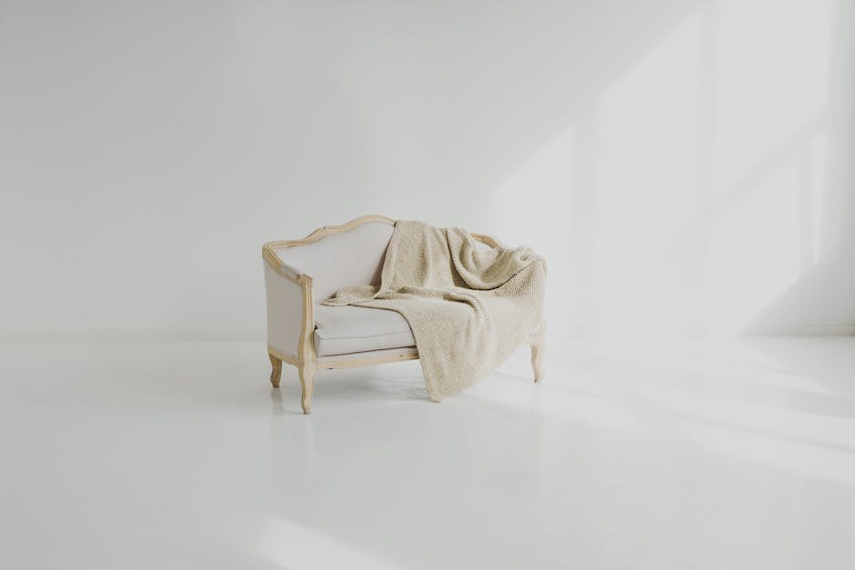 An elegant, vintage-style loveseat with a white upholstered fabric and wooden frame, positioned in a bright, minimalistic room. A beige throw blanket is casually draped over one arm and the backrest of the sofa, which is placed on a smooth, polished white floor. The room features a clean, white wall background and soft natural light spilling in through an unseen window, creating subtle shadows on the floor. The scene appears spotless and well-maintained, reflecting thorough surface cleaning, including furniture and floors, consistent with domestic cleaning and sanitisation standards. This setting exemplifies professional upholstery cleaning and surface maintenance services by Cleaners W6, ensuring a hygienic and pristine environment.