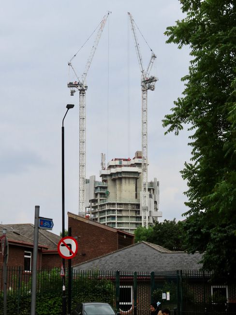 A construction site in Hammersmith with two large tower cranes working on a multi-storey building framework, featuring concrete floors and supporting columns. The cranes are equipped with long booms and counterweights, operating over the partially built structure. In the foreground, residential rooftops, a streetlamp, a no U-turn traffic sign, and green trees are visible, indicating a suburban area. The scene is under overcast skies, with diffused natural lighting highlighting the construction activity. Cleaners W6 offers surface cleaning and deep cleaning services for properties like this, focusing on hygiene and maintenance of surfaces such as glass, tiles, and other materials often involved in construction or post-renovation cleaning.