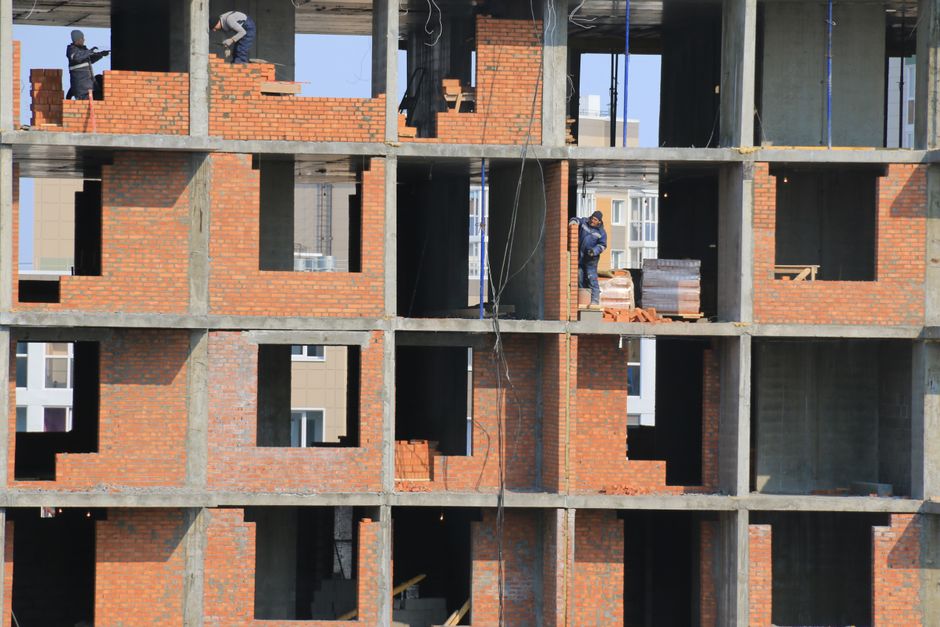 An under-construction residential building with a visible brick façade and exposed concrete structural elements. The building has multiple open window and door frames without glazing, revealing unfinished interiors. Construction workers are present on the different floors; one worker is standing near a stack of bricks on a middle level, wearing a blue jacket and helmet, while another is working on the top floor, dressed in a safety vest. Wooden planks and construction materials are scattered throughout the site. The exterior surfaces are predominantly red brick with some concrete support beams, and natural daylight illuminates the scene. This image illustrates the ongoing building process, highlighting the structural framework typical of modern multi-storey residential developments, aligning with the context of renovation and post-construction cleaning services provided by Cleaners W6.
