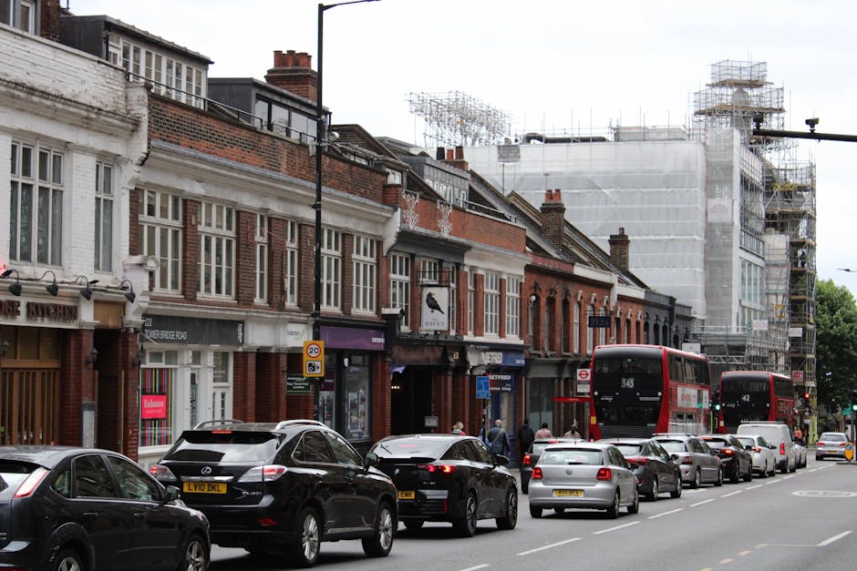 A construction site in Hammersmith with two large tower cranes working on a multi-storey building framework, featuring concrete floors and supporting columns. The cranes are equipped with long booms and counterweights, operating over the partially built structure. In the foreground, residential rooftops, a streetlamp, a no U-turn traffic sign, and green trees are visible, indicating a suburban area. The scene is under overcast skies, with diffused natural lighting highlighting the construction activity. Cleaners W6 offers surface cleaning and deep cleaning services for properties like this, focusing on hygiene and maintenance of surfaces such as glass, tiles, and other materials often involved in construction or post-renovation cleaning.