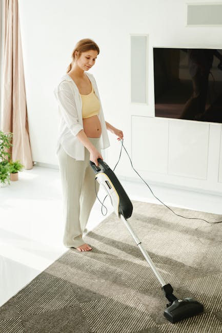 A young woman with light brown hair, dressed in a white cardigan and beige pants, is vacuuming a patterned gray carpet in a bright, modern living room. The room features white walls, a large flat-screen television mounted on a white built-in unit, and natural light coming through beige curtains. She is holding a black and white vacuum cleaner with a long metal tube, actively cleaning the area. The environment appears clean and tidy, with the wooden floor visible near the carpet, and the space is well-lit with soft natural illumination. She is engaged in surface cleaning, carried out by Cleaners W6, providing professional deep cleaning and sanitisation services in the W6 area, as described on the page about same-day cleaning in Hammersmith.