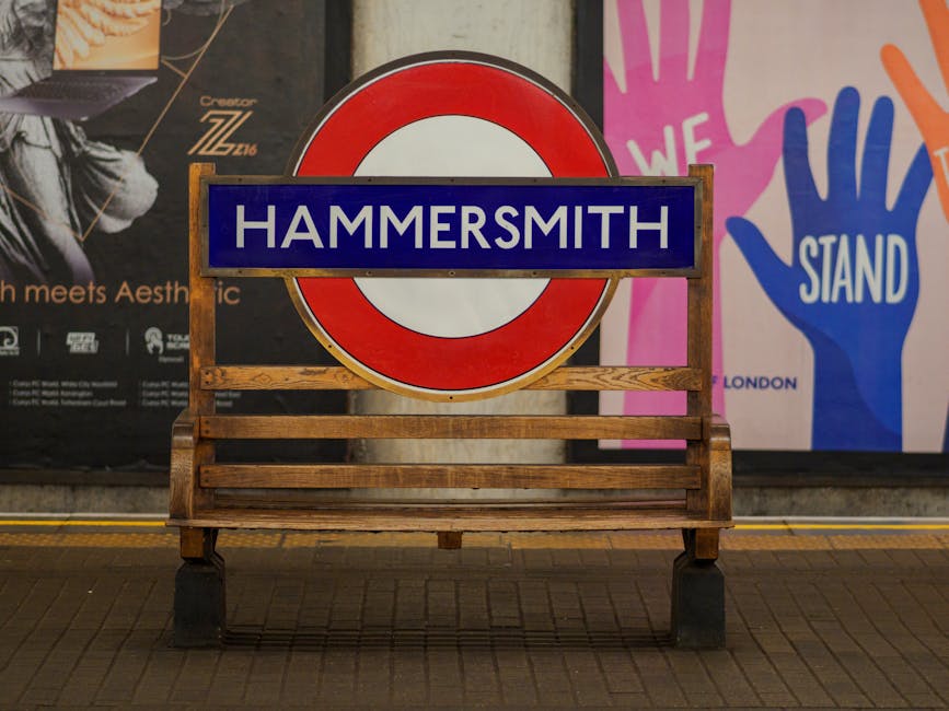 A wooden bench with horizontal slats is positioned in front of a large, circular London Underground sign that reads 'HAMMERSMITH' on a black background with a red and white logo. Behind the sign, there are posters on a wall, including one with colorful hand illustrations and the word 'STAND' in bold white letters. The area appears clean and well-lit, with no visible dust or dirt on the surfaces. The floor is made of dark tiles, and the lighting highlights the shiny, smooth surfaces of the sign and the polished appearance of the surrounding environment, suggesting a well-maintained space suitable for surface cleaning and hygiene maintenance, as provided by Cleaners W6.